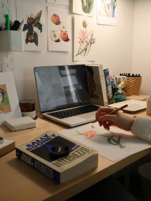 Artist designing watercolor flowers at a creative indoor workspace with books and laptop.
