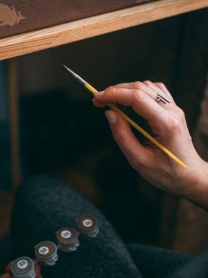 Close-up of an artist's hand holding a paintbrush while painting on canvas indoors.