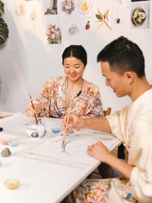 Two adults engaging in a Japanese art class, painting with brushes in a bright studio.